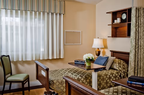 A nursing center room with two single beds featuring patterned bedspreads, a small table with a lamp and a flower vase, a chair near a window with sheer curtains, and a wall shelf with a clock and decorative items.