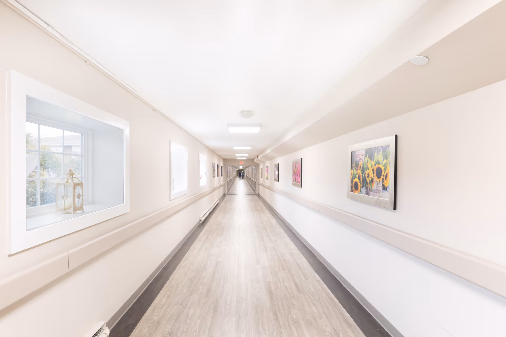 A long, bright hallway in a nursing and rehabilitation facility with light-colored walls and wood-patterned flooring. The hallway has handrails on both sides and framed pictures hanging on the right wall. There are windows on the left side letting in natural light, and ceiling lights illuminate the corridor.