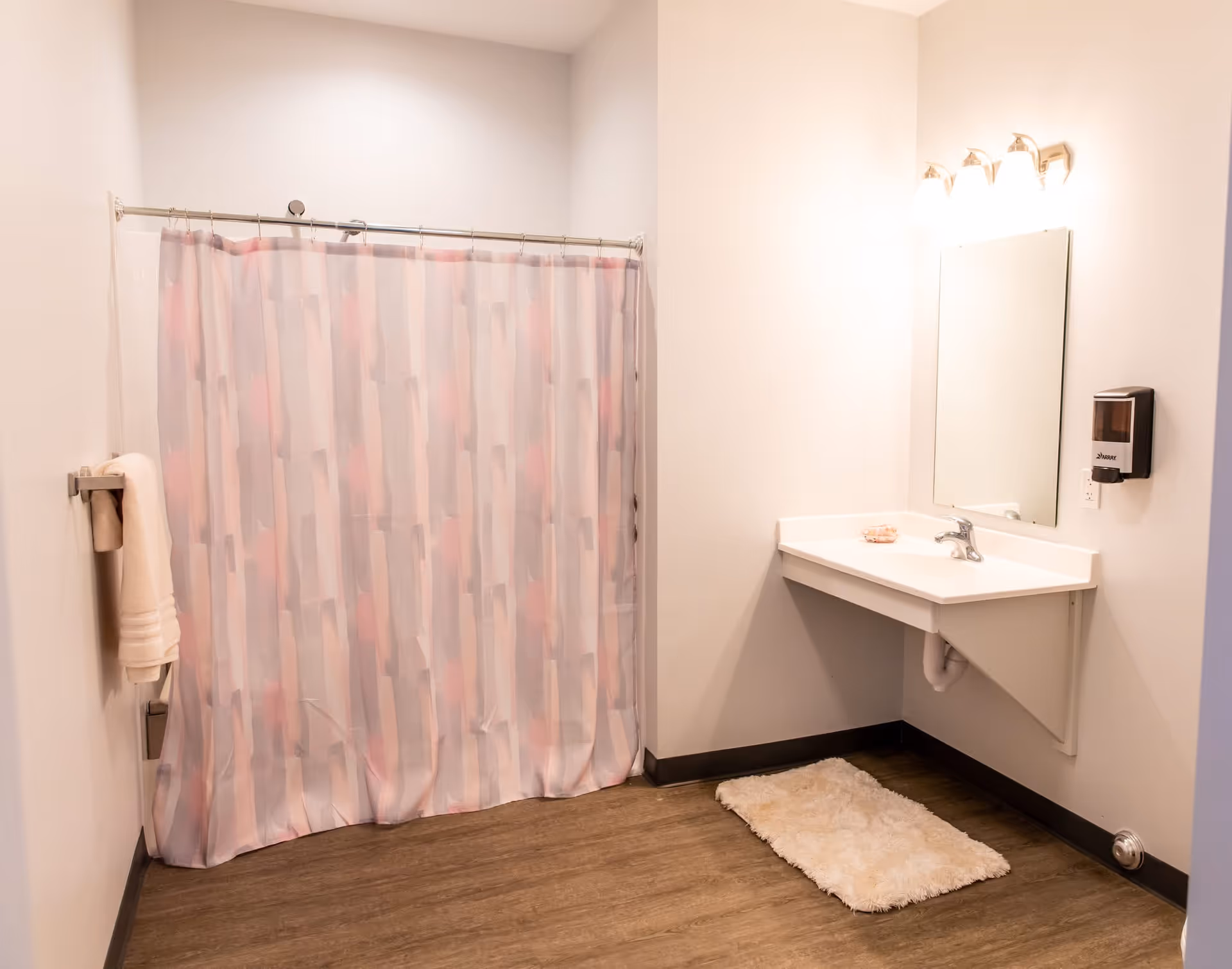 A clean bathroom with a shower area covered by a patterned shower curtain, a wall-mounted sink with a mirror above it, a soap dispenser on the wall, a towel hanging on a rack, and a fluffy white bath mat on the wooden floor.