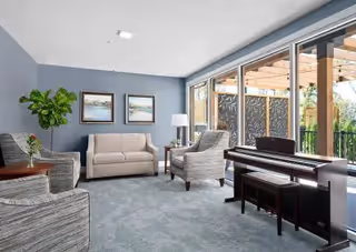 Bright community living room with a sofa, armchairs, a piano, and floor-to-ceiling glass doors opening to a pergola-covered outdoor area.