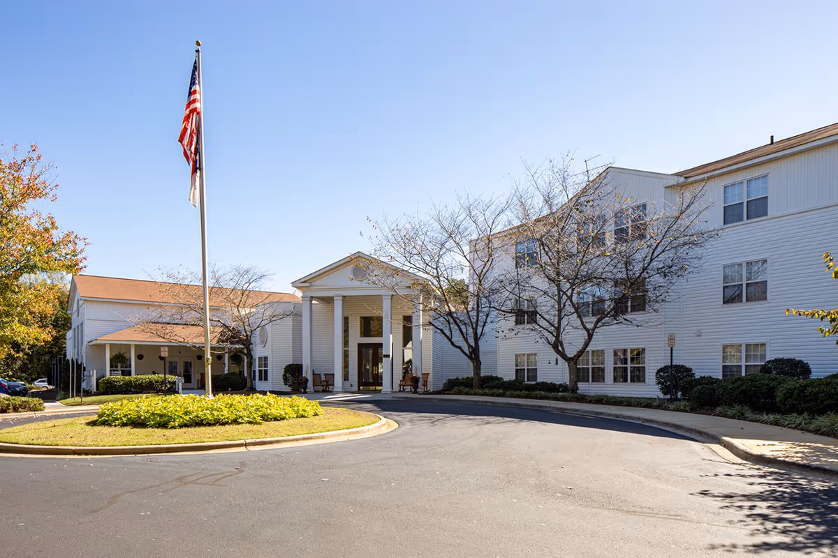White multi-story senior living building with a columned entrance, circular driveway and an American flag on a flagpole.