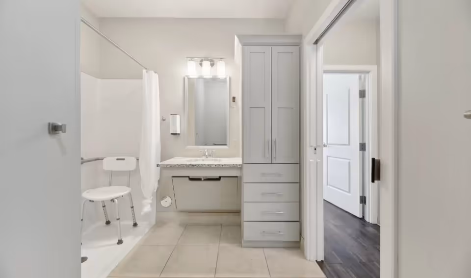 A clean, modern bathroom with a walk-in shower featuring a white shower chair and a white shower curtain. There is a vanity with a granite countertop, a mirror with three lights above it, and a tall cabinet with drawers and doors. The bathroom floor is tiled, and an open doorway leads to a room with dark wood flooring.