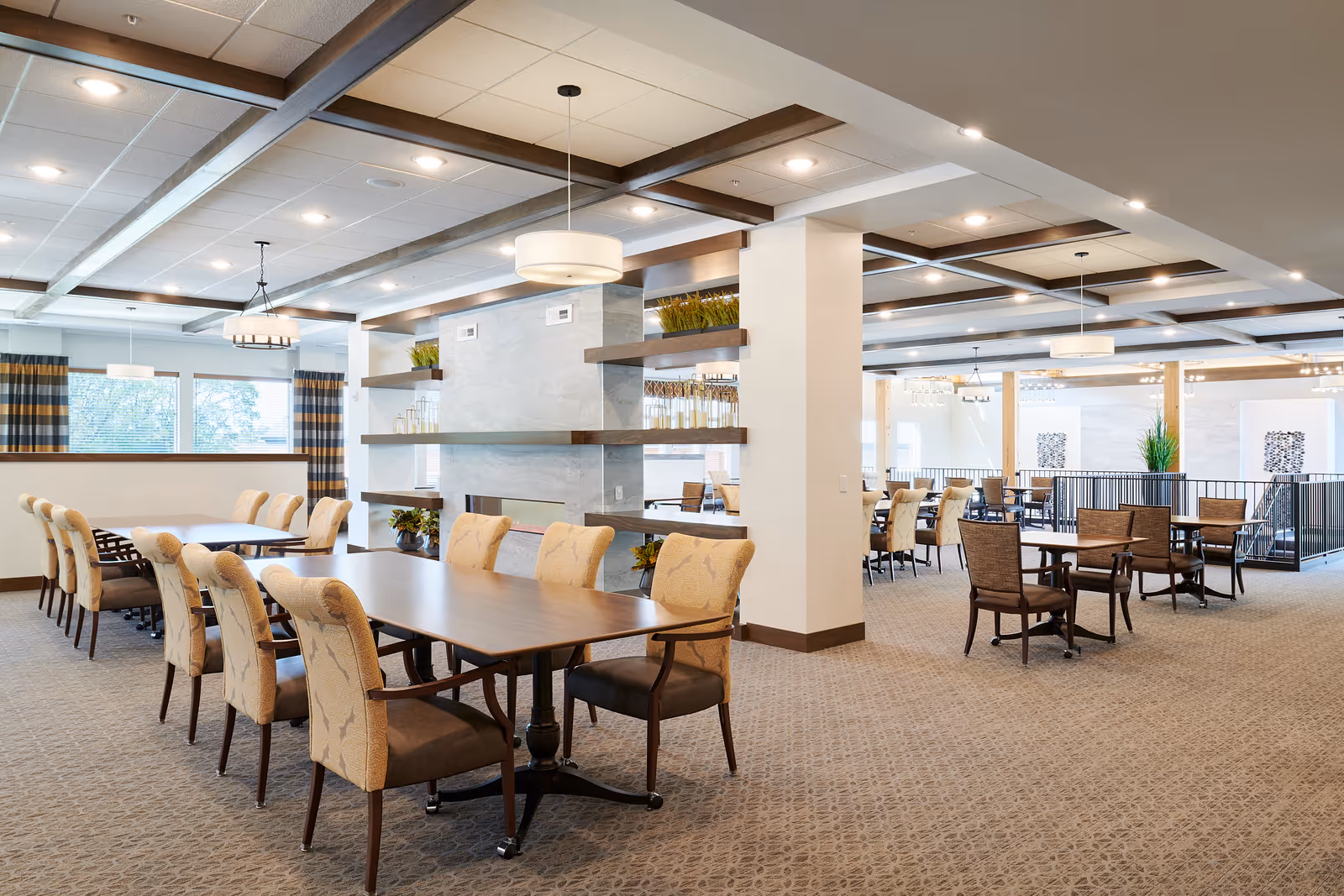 A spacious and well-lit dining area with multiple tables and cushioned chairs arranged neatly. The room features a modern ceiling design with wooden beams and pendant lights. Large windows with striped curtains allow natural light to enter, and decorative shelves with plants and glassware add a touch of elegance.