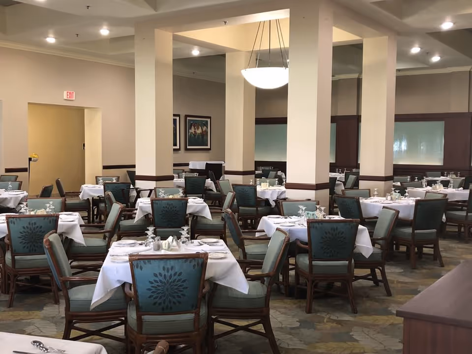A spacious dining room with multiple tables covered in white tablecloths, each set with glassware, plates, and silverware. The chairs have wooden frames and teal upholstery with a floral pattern. The room has beige walls, several large pillars, and framed artwork on the walls. Ceiling lights provide illumination.
