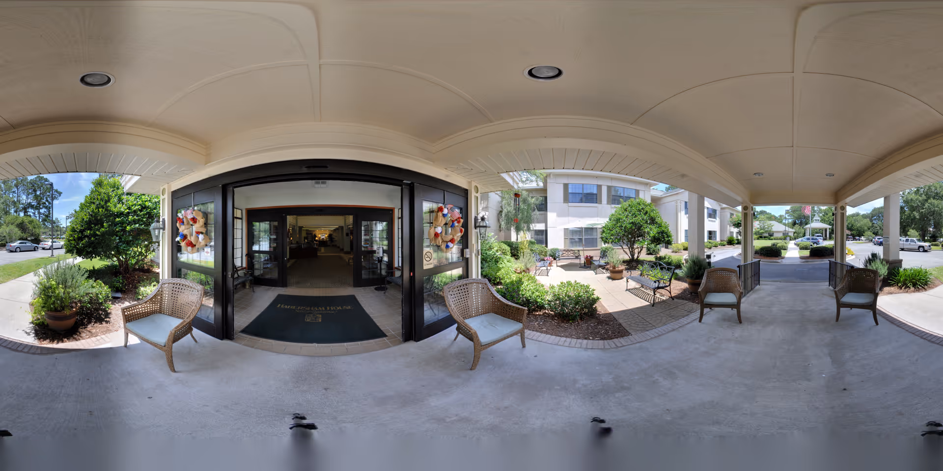 Covered entrance/portico of a senior living facility with wicker chairs, potted plants, benches, and glass double doors leading inside.