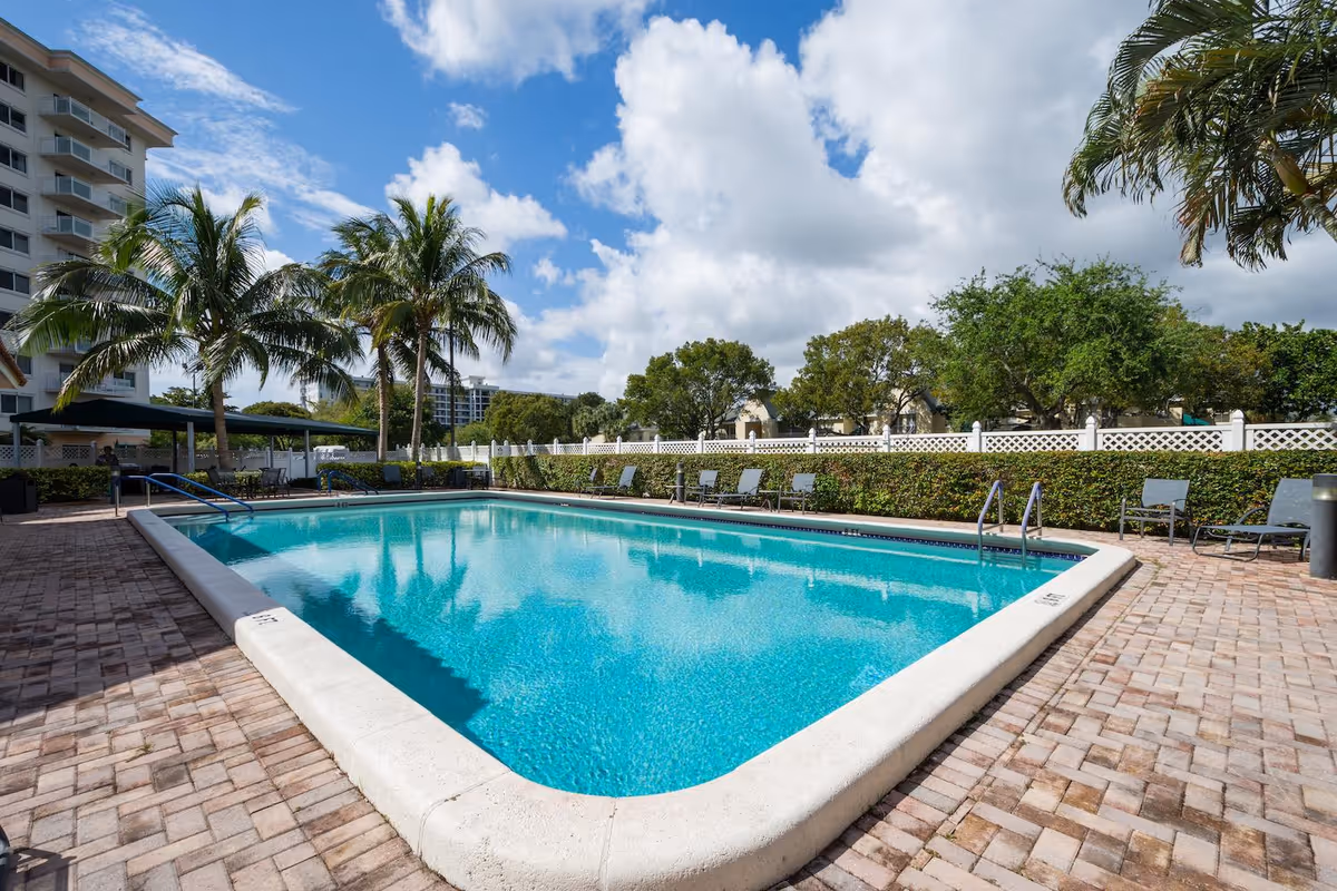 Outdoor swimming pool surrounded by palm trees, lounge chairs, and a paved deck area under a partly cloudy blue sky.