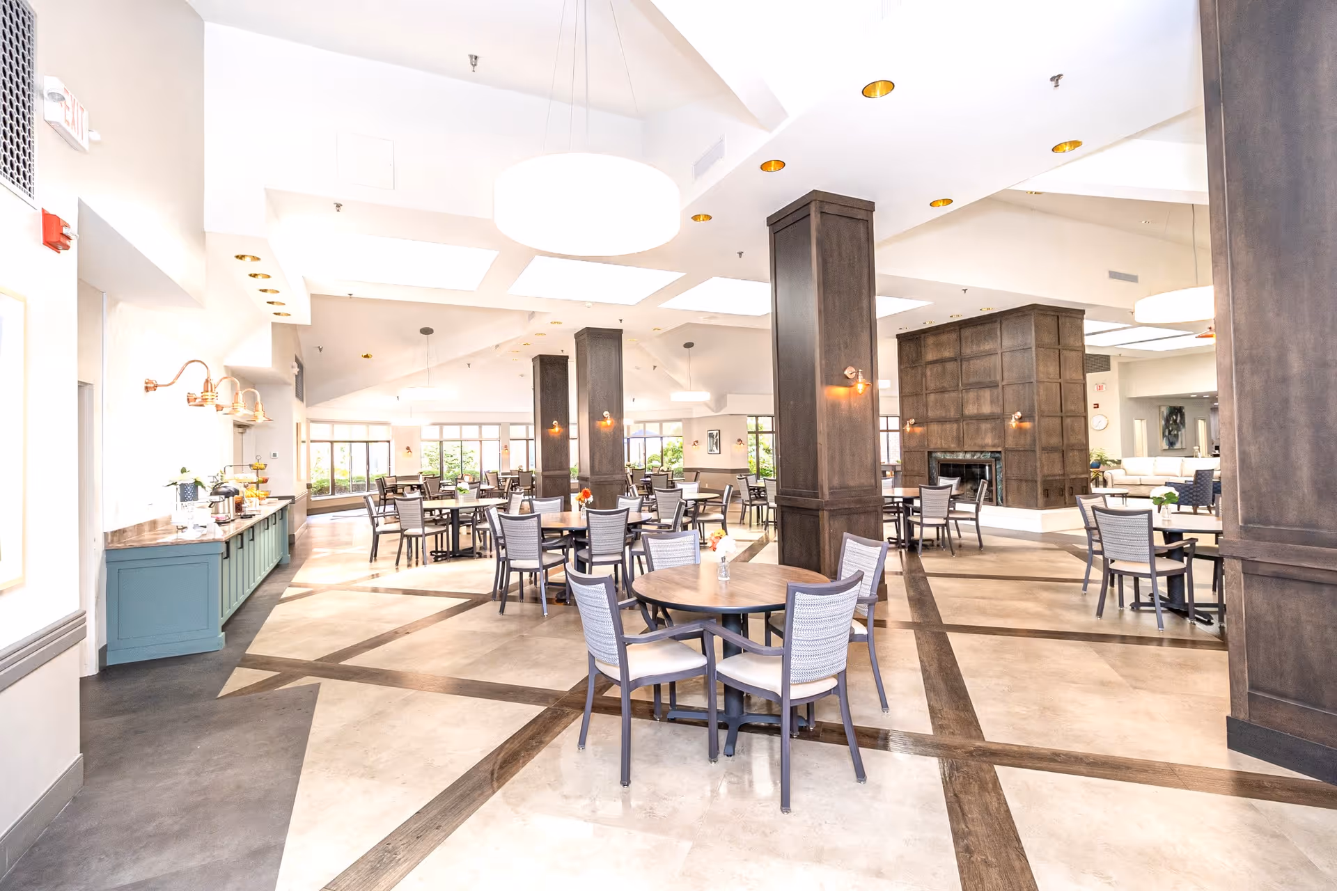 Spacious, well-lit dining room with round tables and chairs, tall wooden pillars, and a buffet counter.