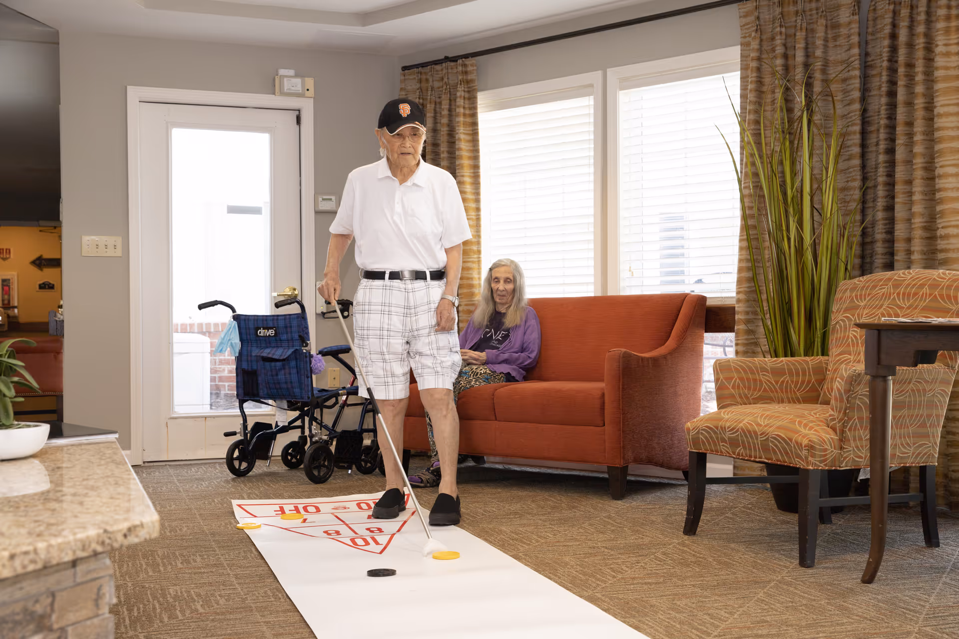 An elderly man wearing a white shirt, plaid shorts, and a black cap is playing shuffleboard indoors on a white shuffleboard mat. A woman with long gray hair, wearing a purple top, is sitting on an orange couch in the background. There is a blue walker near the door, a patterned armchair, a tall green plant, and large windows with blinds behind the couch.