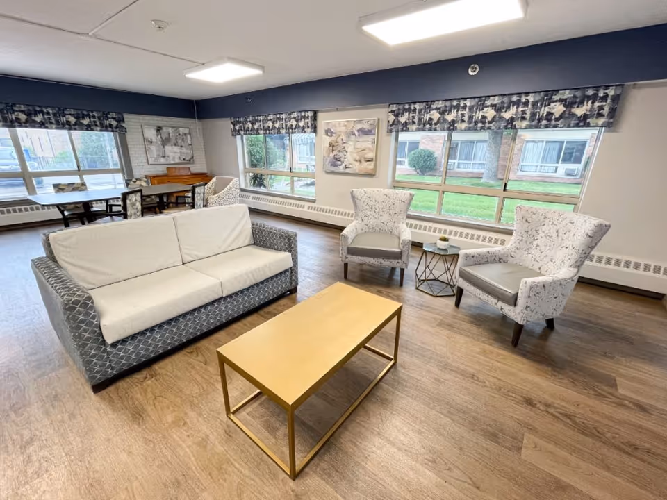 A bright and spacious living room area with a modern sofa, two patterned armchairs, a gold-colored rectangular coffee table, and a small side table with a plant. Large windows with patterned valances let in natural light and offer a view of green grass and bushes outside. In the background, there is a dining table with chairs and a piano against the wall.