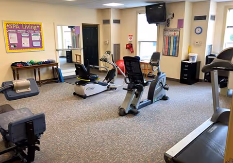 A fitness room in a senior living facility with exercise equipment including stationary bikes, a treadmill, and weights. The room has beige walls, carpeted floor, a wall-mounted TV, a bulletin board labeled 'SPA Living', and a window letting in natural light.