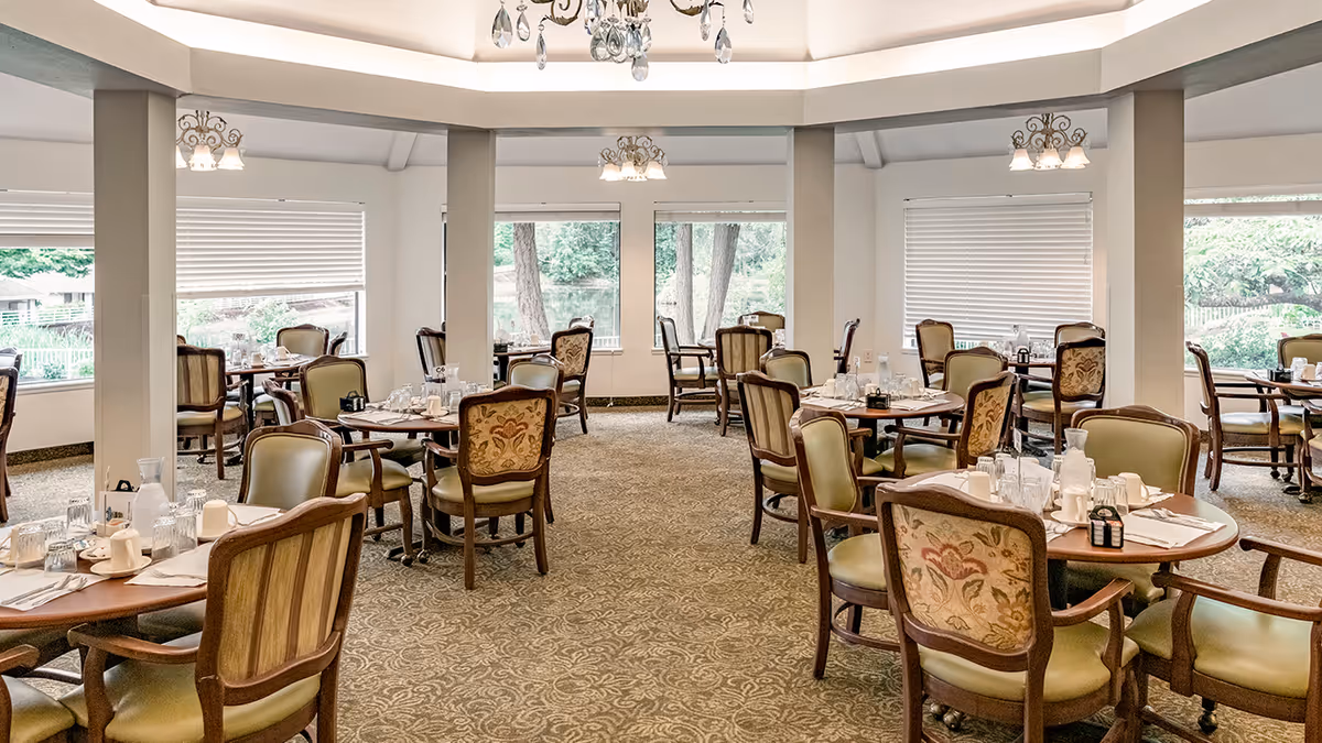 A spacious dining room with multiple round wooden tables set with glasses, cups, and napkins. The chairs have cushioned seats and backs with floral upholstery. Large windows with white blinds allow natural light to fill the room, and there are decorative light fixtures hanging from the ceiling.