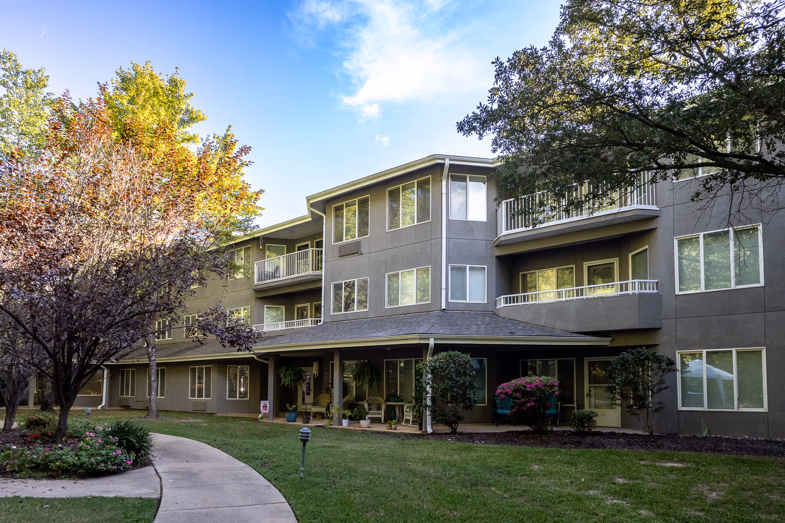 Three-story senior living building with balconies, a covered entrance, landscaped lawn and a curved walkway.