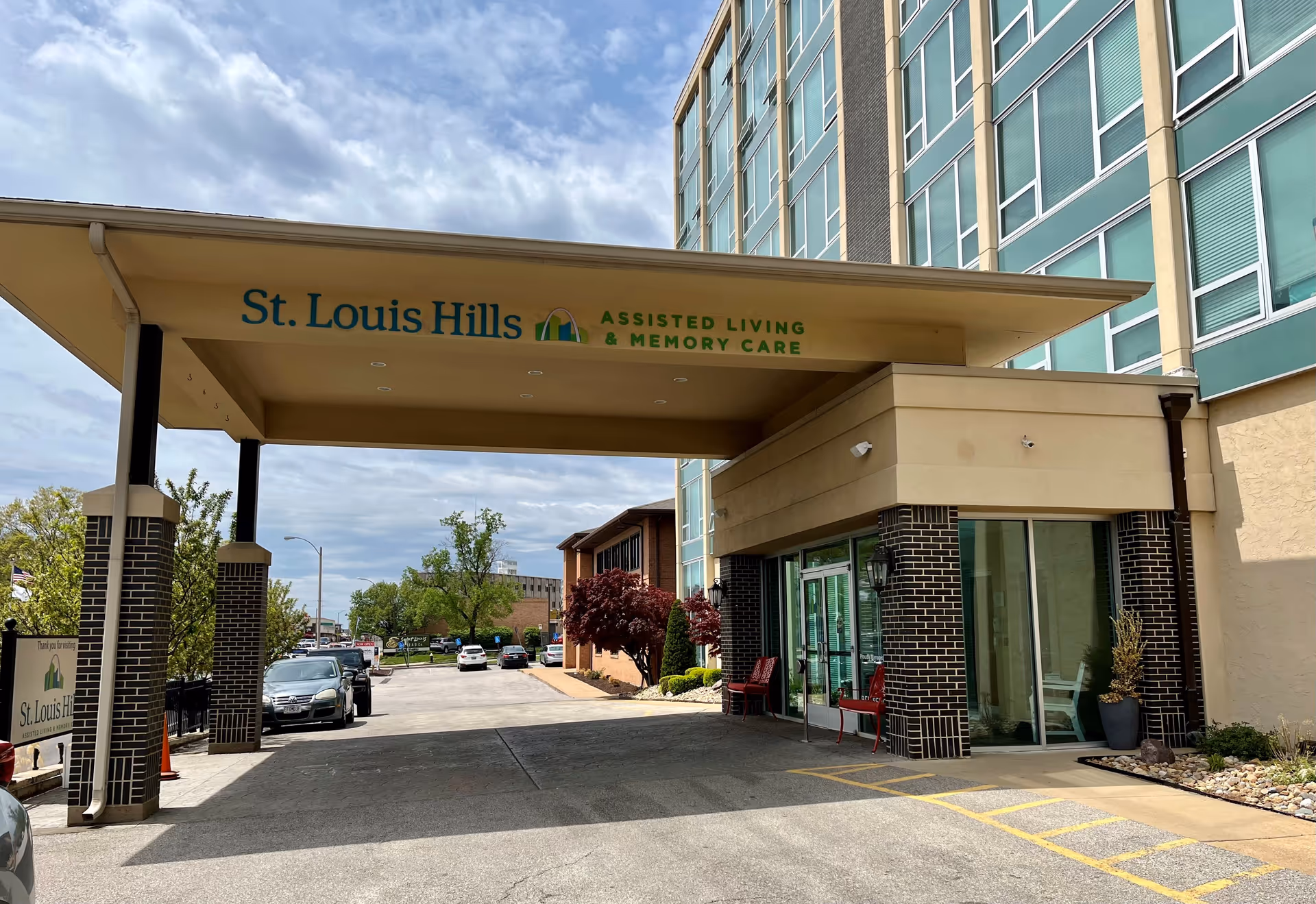 Entrance to St. Louis Hills Assisted Living & Memory Care facility with a covered drop-off area, glass doors, and a multi-story building in the background. Several cars are parked along the driveway and trees with green and red leaves are visible.