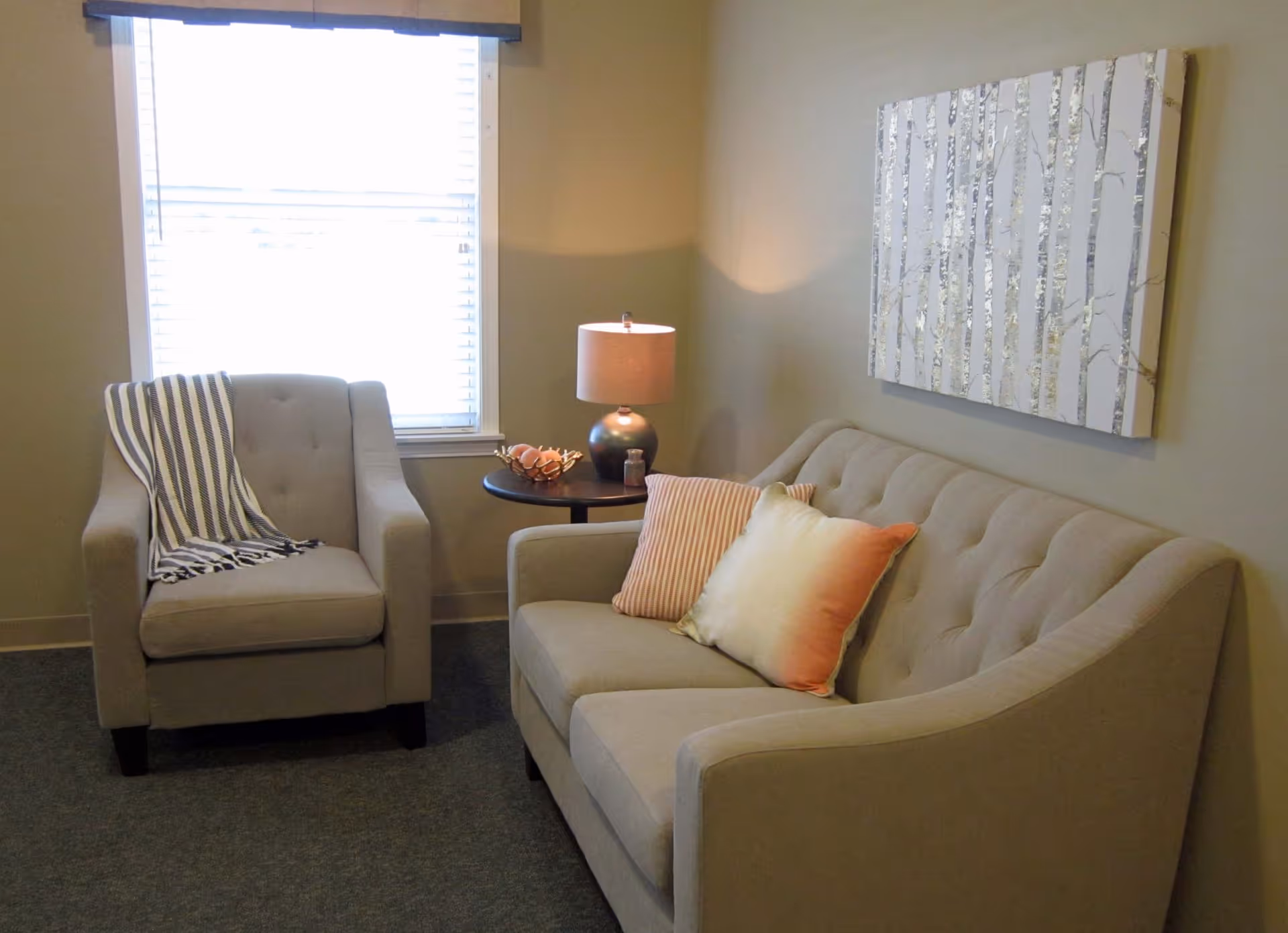 A cozy living room area with a beige armchair draped with a striped blanket, a matching beige sofa with two decorative pillows, a round side table with a lamp and decorative items, and a window with blinds letting in natural light. A painting of birch trees hangs on the wall above the sofa.