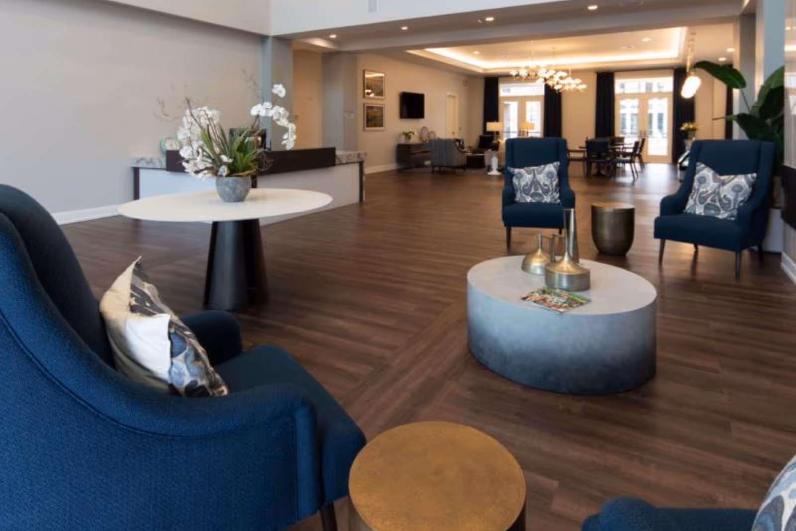 A spacious senior living facility common area with dark wood flooring, blue upholstered armchairs with patterned pillows, a round white table with a flower arrangement, a gray oval coffee table with decorative items, and a dining area with a chandelier in the background.