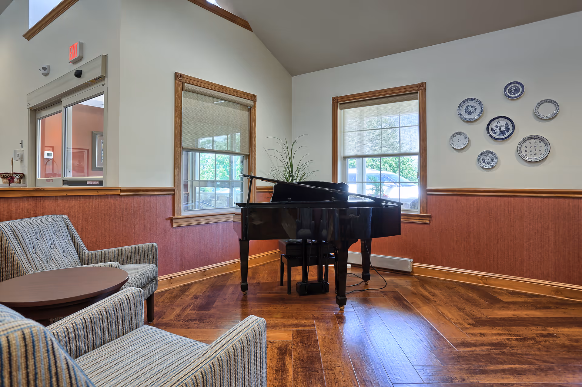 A bright lounge with a black grand piano, two upholstered chairs and a small round table, windows, and decorative plates on the wall.