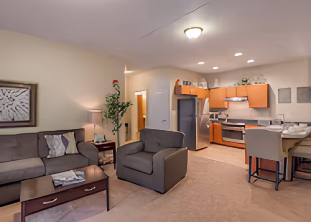 Interior view of a senior living facility showing a living room area with a gray sofa, armchair, coffee table, side table with a lamp, and a plant. In the background, there is an open kitchen with wooden cabinets, stainless steel refrigerator, stove, and a small dining area with chairs and a table.