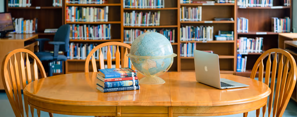 A wooden table with four matching wooden chairs around it in a library setting. On the table, there is a globe, a stack of books, and an open laptop. Behind the table, there are bookshelves filled with books and a computer workstation with a chair.