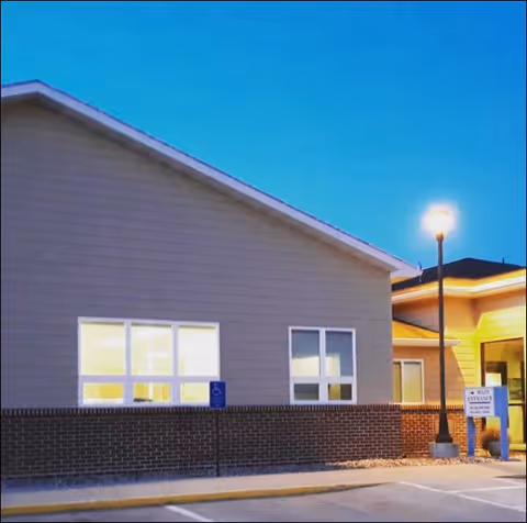 Exterior view of a building at dusk with illuminated windows, a street lamp, and a sign indicating the main entrance. The building has beige siding with a brick lower section and a parking area in front.