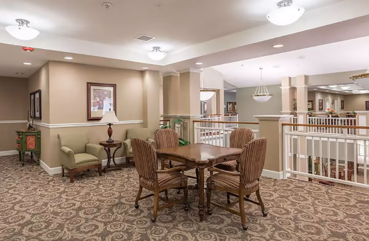 Interior common area of Regency Pullman featuring a wooden table with four striped upholstered chairs on a patterned carpet. There are two armchairs with a small round table and lamp against a beige wall with framed artwork. The space is well-lit with ceiling lights and has white railings overlooking a lower level.