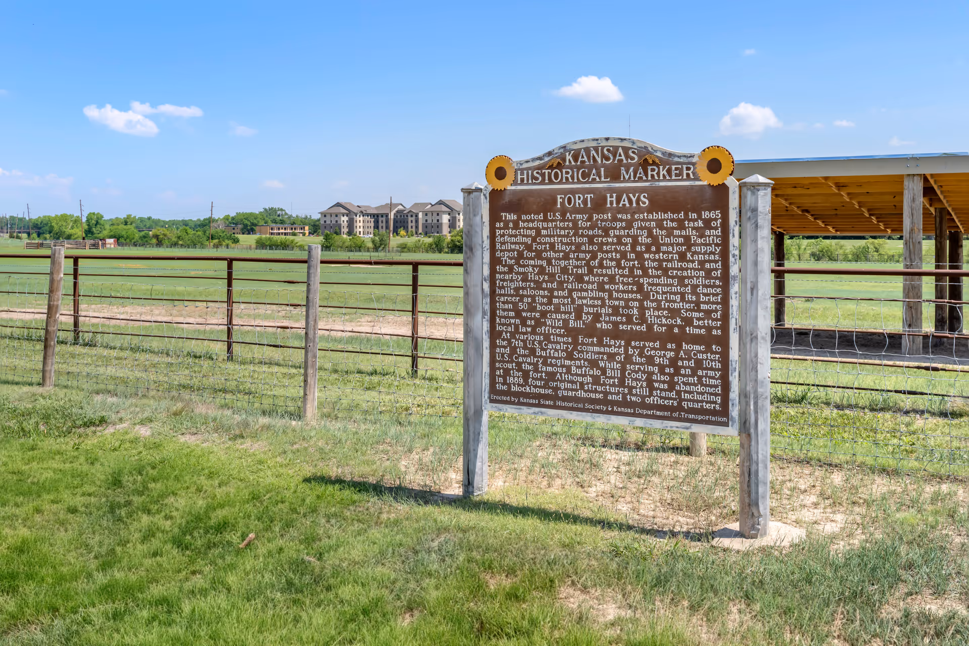A Kansas Historical Marker sign for Fort Hays stands in a grassy area near a wooden fence. In the background, there is a large building complex and a covered structure, under a blue sky with a few clouds.
