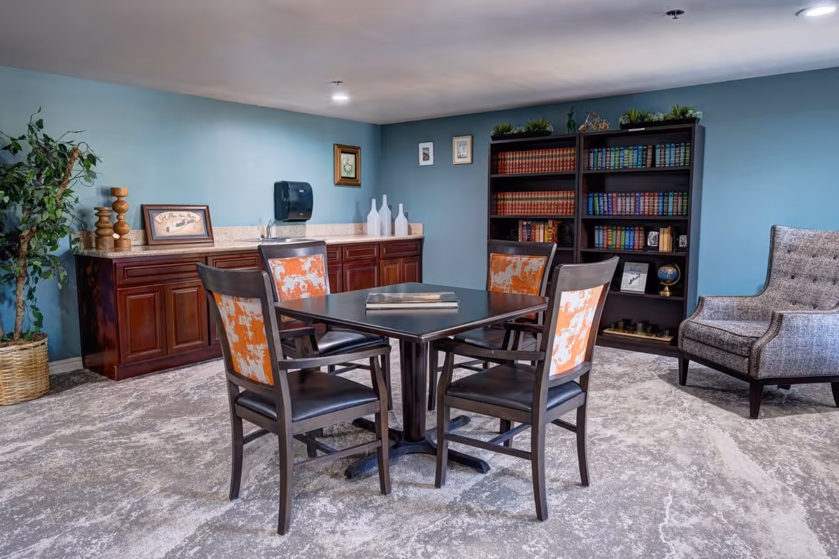 A cozy interior room with a square table surrounded by four chairs with orange and white patterned backs. Behind the table is a wooden cabinet with decorative items and a paper towel dispenser mounted on the wall. To the right, there is a tall bookshelf filled with books and decorative plants on top, and a gray upholstered armchair with nailhead trim. The walls are painted light blue and the floor is covered with a patterned carpet.
