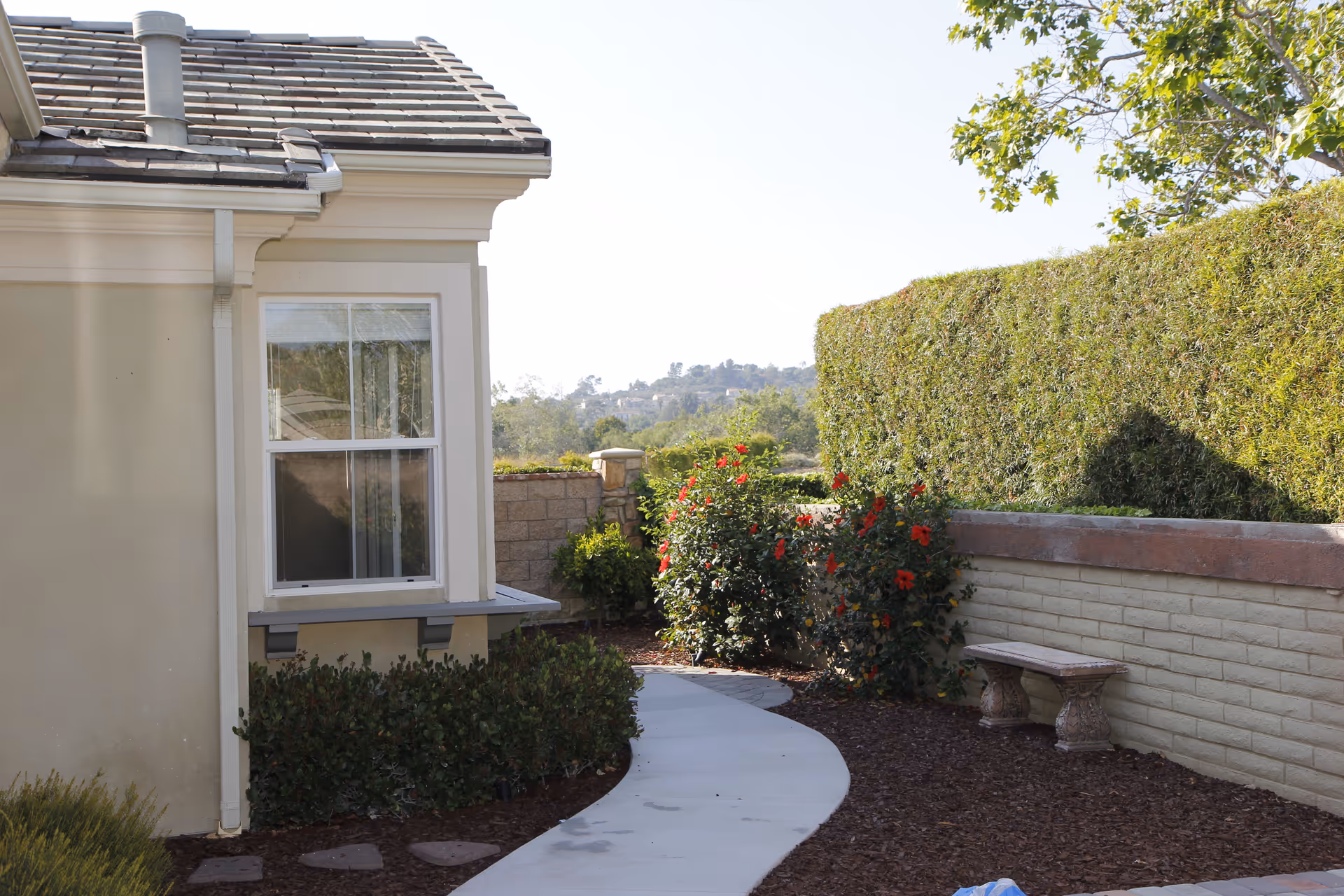 A curved concrete pathway runs alongside the exterior wall of a beige building with a window. On the right side of the path, there is a low brick wall with a stone bench and a tall hedge behind it. Flowering bushes with red flowers are planted near the wall, and trees and hills are visible in the background under a clear sky.