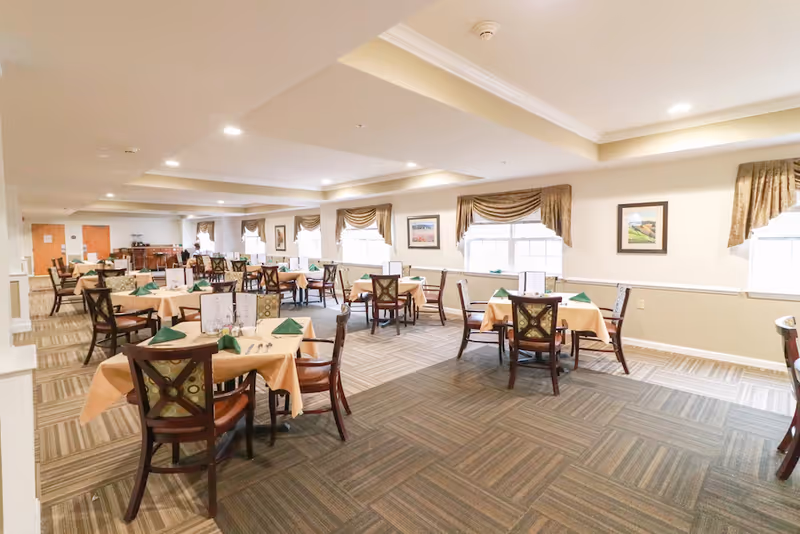 A spacious dining room with multiple tables covered in beige tablecloths and set with green napkins and silverware. The room has large windows with brown valances, framed artwork on the walls, and a carpeted floor with a patterned design. The ceiling features recessed lighting and a tray ceiling design.
