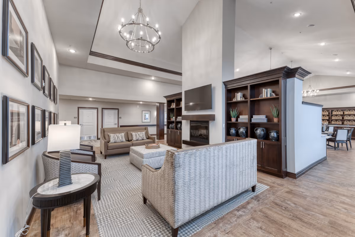 A spacious senior living facility common area with comfortable seating including a beige sofa, a patterned armchair, and a large ottoman. The room features a modern fireplace with a mounted flat-screen TV above it, flanked by dark wood built-in bookshelves decorated with books, plants, and vases. The walls are adorned with framed pictures, and a round chandelier hangs from the high ceiling. The flooring is a combination of wood and patterned carpet, and there is a dining area visible in the background.