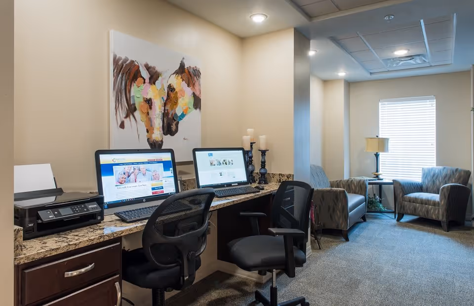 A small office or computer workstation area in a senior living facility with two desktop computers, a printer, two black office chairs, and a colorful painting of two horses on the wall. In the background, there is a seating area with two patterned armchairs, a small table with a lamp, and a window with blinds.