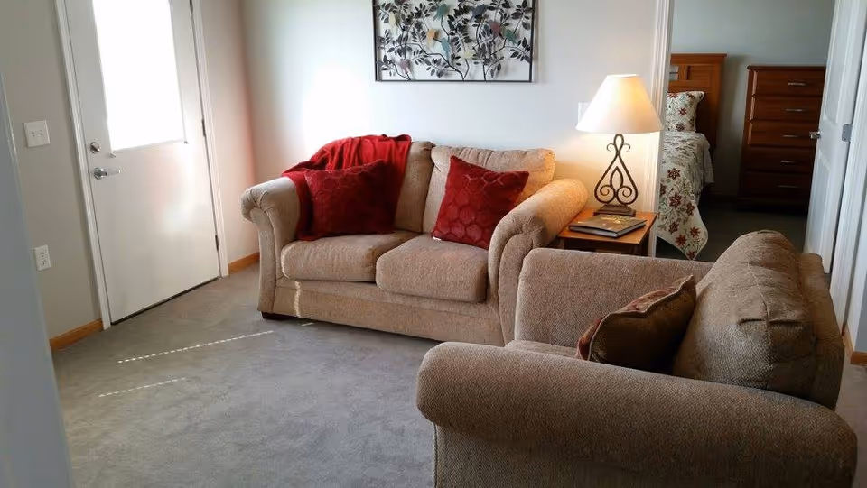 A cozy living room area with two beige sofas, one with red cushions and a red throw blanket. A side table with a decorative lamp and a book is placed between the sofas. In the background, a bedroom with a bed covered in a floral bedspread and a wooden dresser is visible. A door with a window is on the left side of the room.