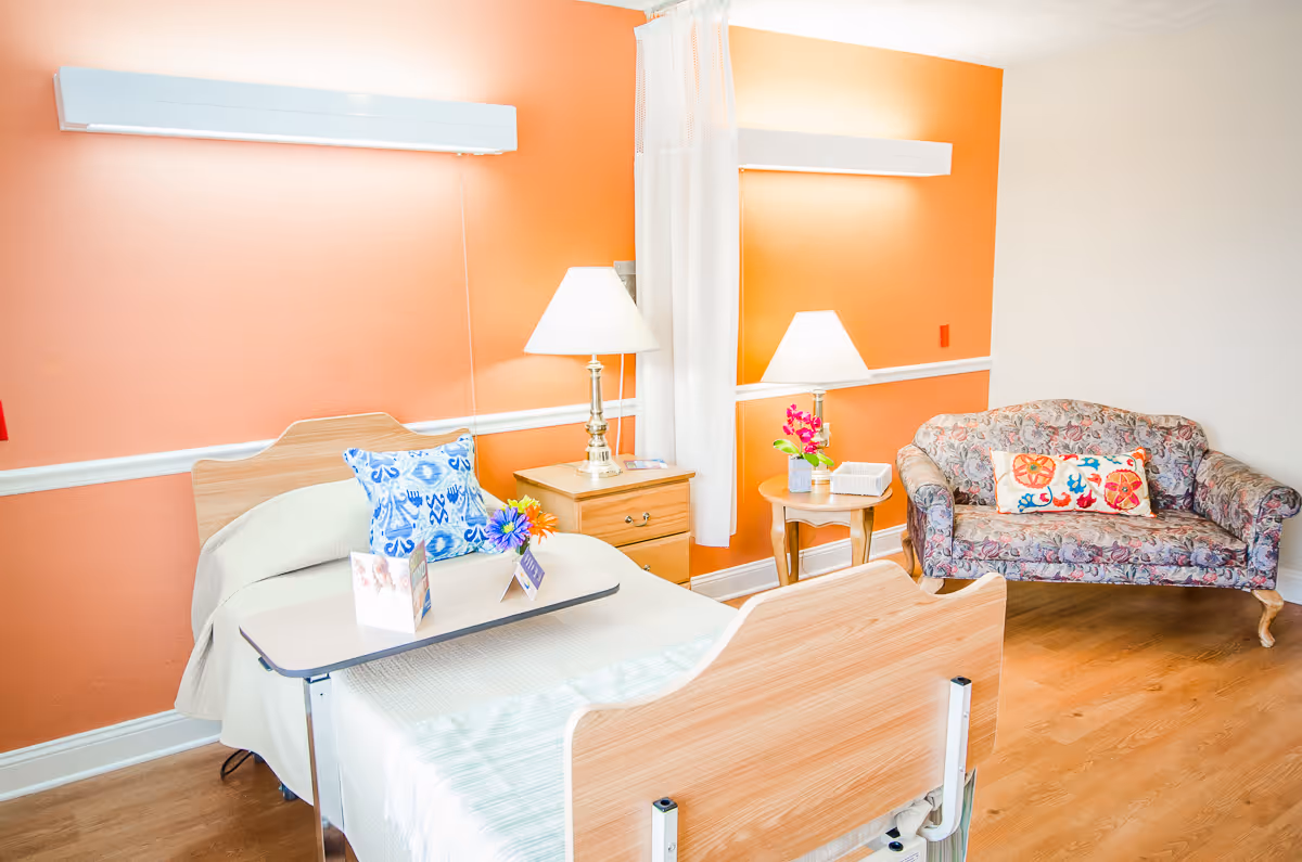 A bright and clean senior living bedroom with an adjustable hospital-style bed featuring a blue patterned pillow and a small table with flowers and cards. The room has orange and white walls, two bedside tables with lamps, and a floral-patterned loveseat with a colorful decorative pillow on the right side.