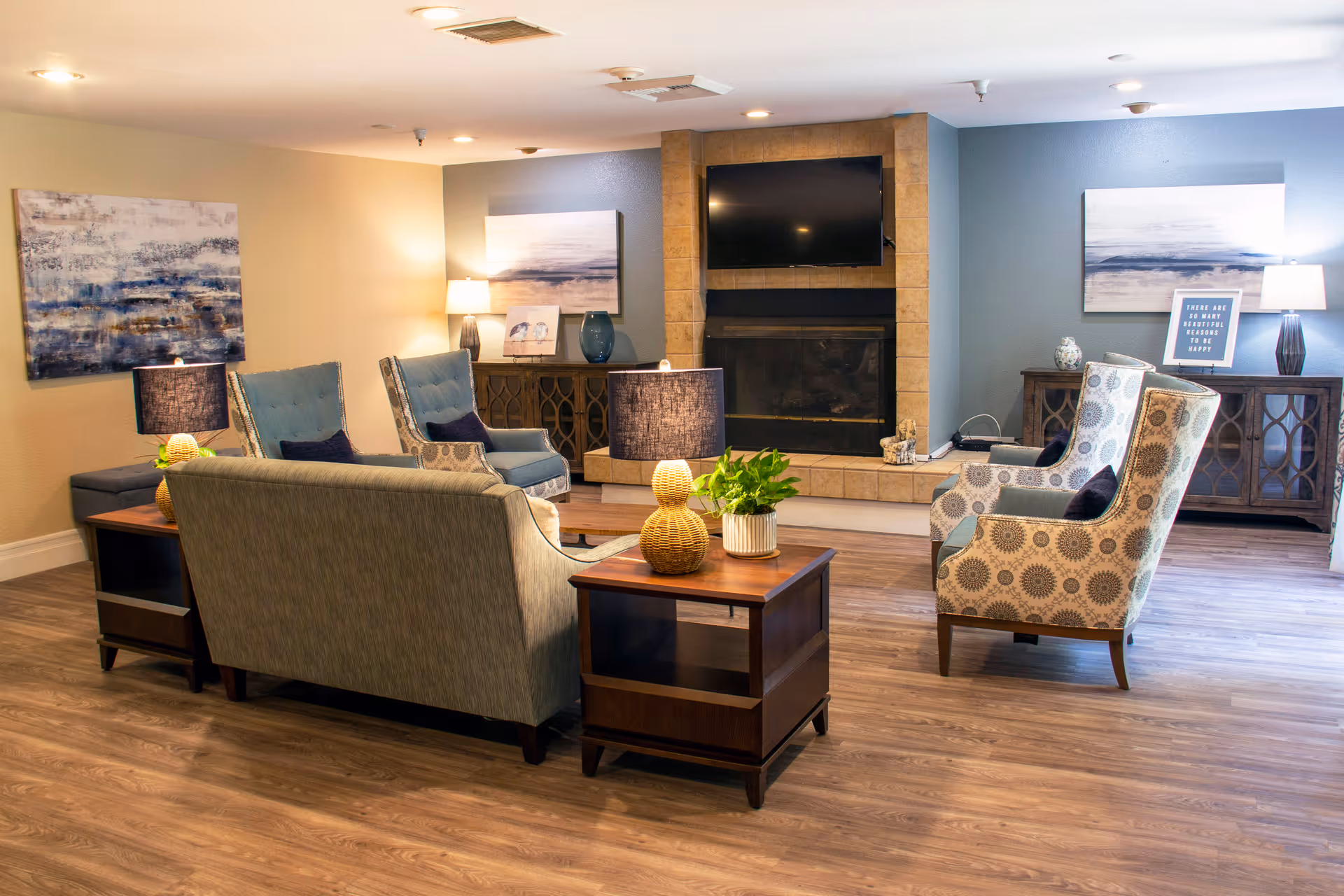 A cozy living room area with a sofa, two patterned armchairs, and two blue armchairs arranged around a fireplace with a mounted flat-screen TV above it. The room has wooden flooring, two side tables with lamps and plants, and abstract artwork on the walls.