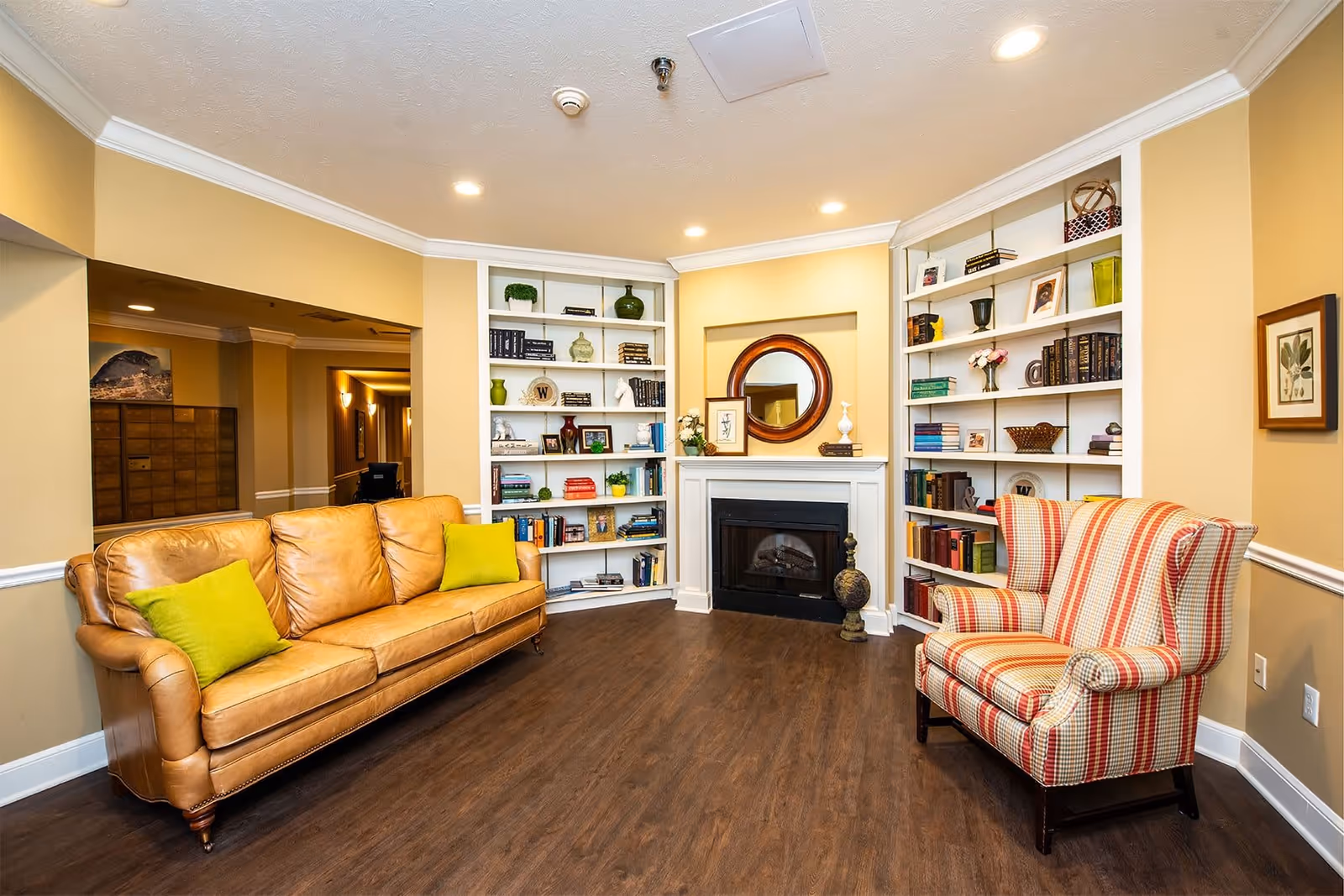 A cozy living room area with a tan leather sofa adorned with green pillows on the left and a red and beige striped armchair on the right. The room features built-in white bookshelves filled with books and decorative items on either side of a white fireplace with a round mirror above it. The walls are painted a warm beige color and the floor is dark wood. Recessed ceiling lights illuminate the space.