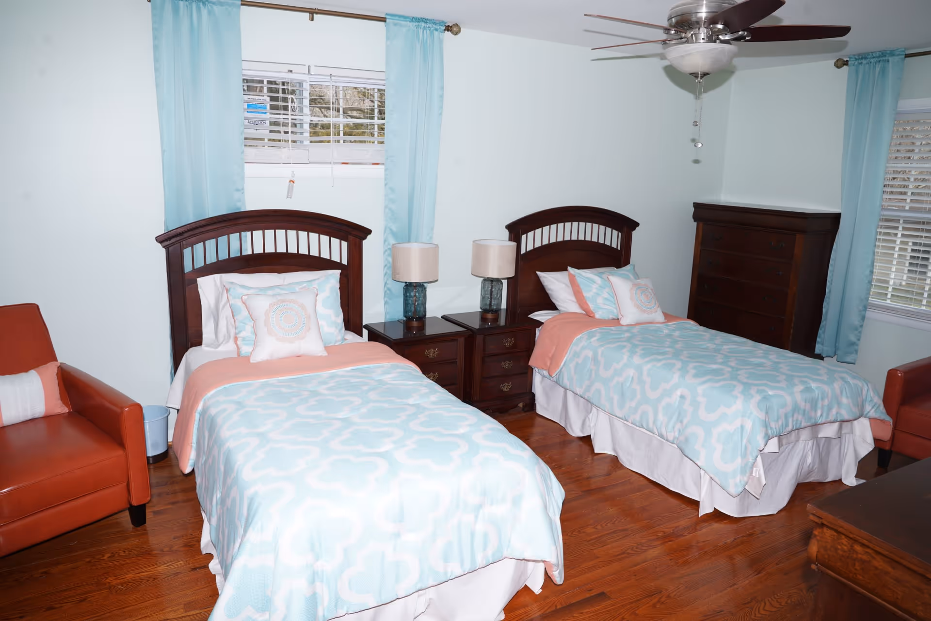 A bedroom with two twin beds featuring wooden headboards, light blue and white patterned bedding with coral accents, and decorative pillows. Between the beds is a dark wooden nightstand with two matching lamps. The room has light blue walls, hardwood floors, two windows with light blue curtains, a ceiling fan with light, a dark wooden dresser, and two coral-colored armchairs.