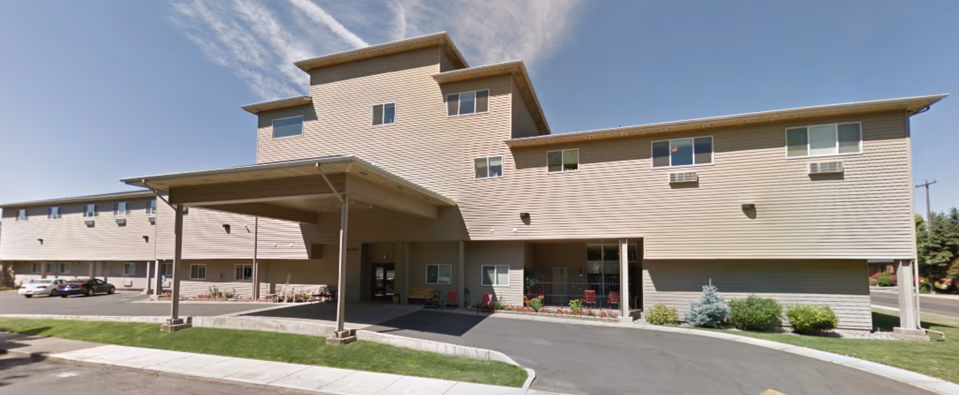 Exterior view of a multi-story assisted living facility building with beige siding, several windows, and a covered entrance area. There are a few cars parked in the parking lot and some landscaping with bushes and grass around the building.