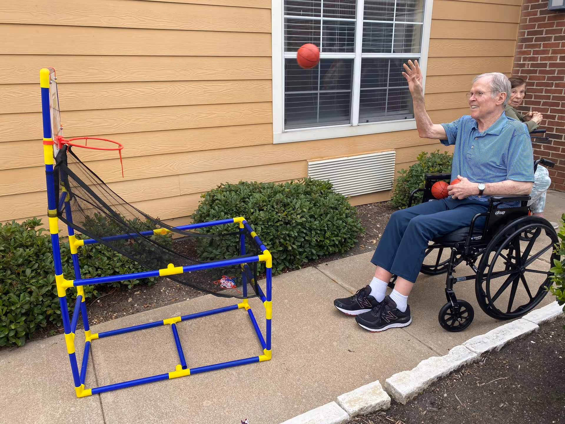 An elderly man in a wheelchair is playing a basketball tossing game outside near a building with beige siding and a window. He is throwing a small red basketball towards a blue and yellow basketball hoop with a netted frame. Another elderly person is seated in the background.