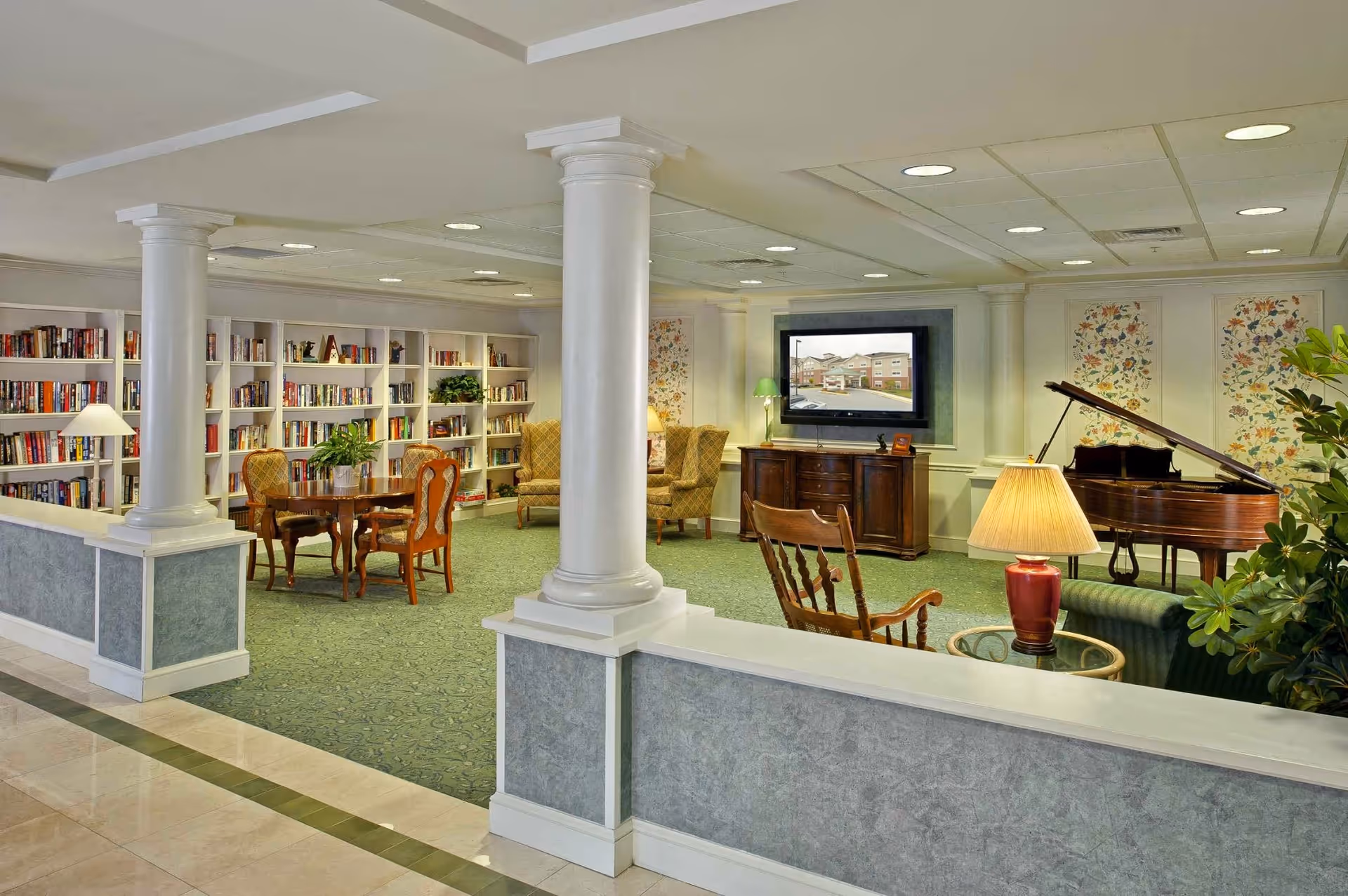 A spacious and well-lit common area in a senior living facility featuring a green carpet, white columns, bookshelves filled with books, a wooden table with chairs, two armchairs, a flat-screen TV mounted on the wall, a grand piano, and several lamps providing warm lighting.