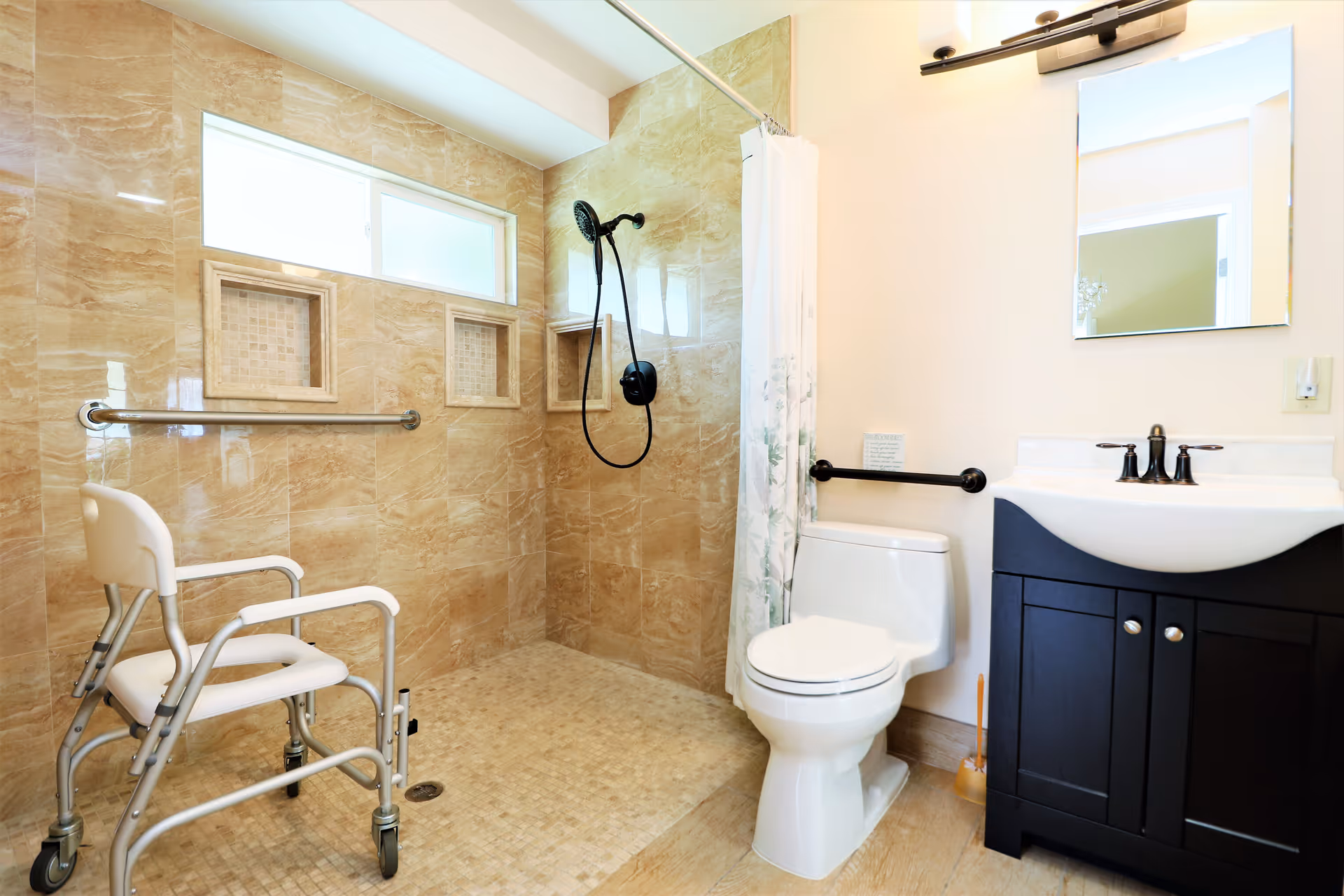 A bright and accessible bathroom featuring a walk-in shower with beige tiled walls and floor, a white shower chair with wheels, a white toilet with a black grab bar beside it, and a dark wood vanity with a white sink and mirror above it.