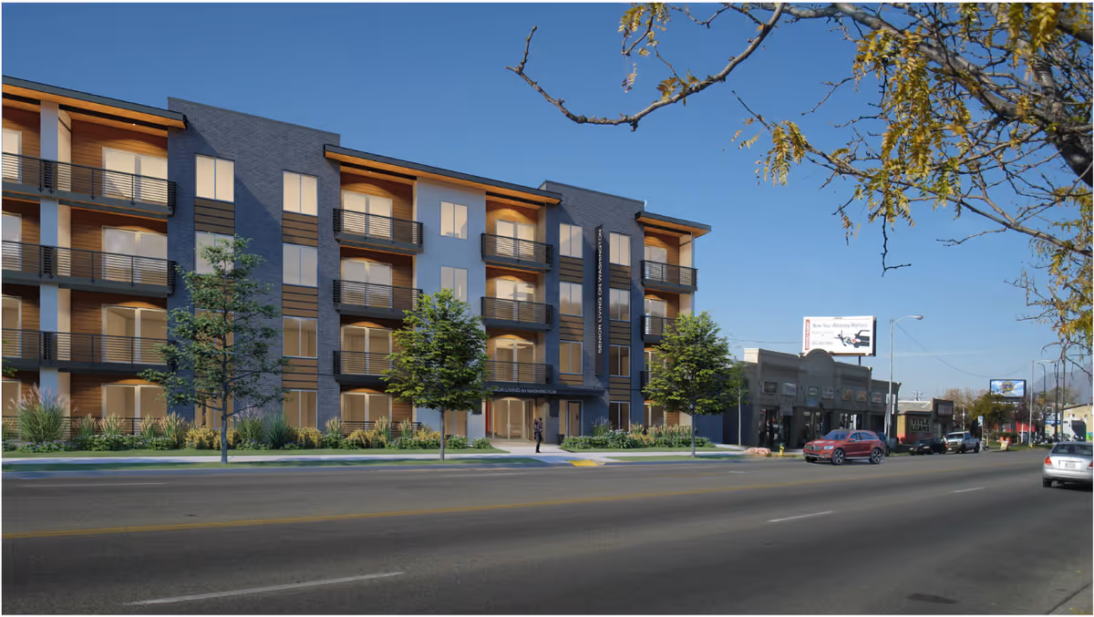 Street view of a modern four-story senior living facility named Senior Living on Washington, with balconies and large windows. The building is surrounded by small trees and landscaping, with a clear blue sky overhead. Nearby are other commercial buildings and parked cars along the road.