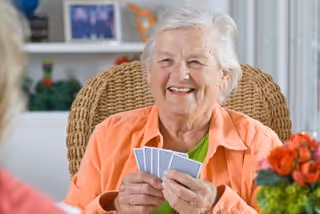 An elderly woman with white hair wearing an orange shirt is smiling and holding playing cards while sitting in a wicker chair. There is a blurred person in the foreground and a colorful flower arrangement on the right side. The background shows a shelf with framed photos and decorative items.