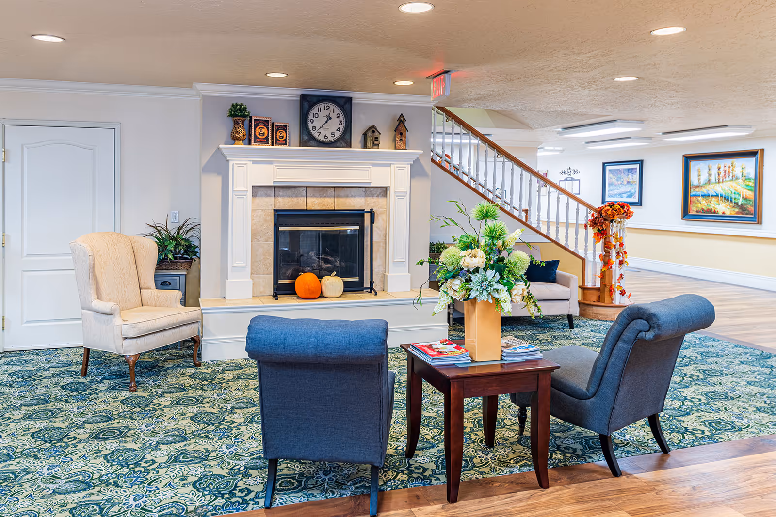 A cozy common room with a fireplace, chairs and a small table topped by a floral arrangement, and a staircase in the background.