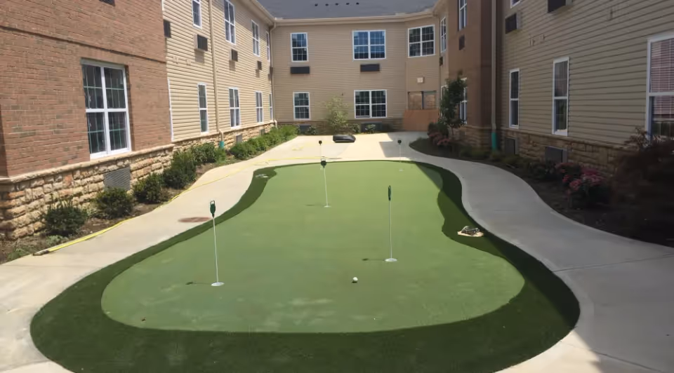 Outdoor courtyard area with a small putting green surrounded by a concrete walkway, bordered by a multi-story building with beige siding and brick accents, and some landscaping with bushes and flowers.