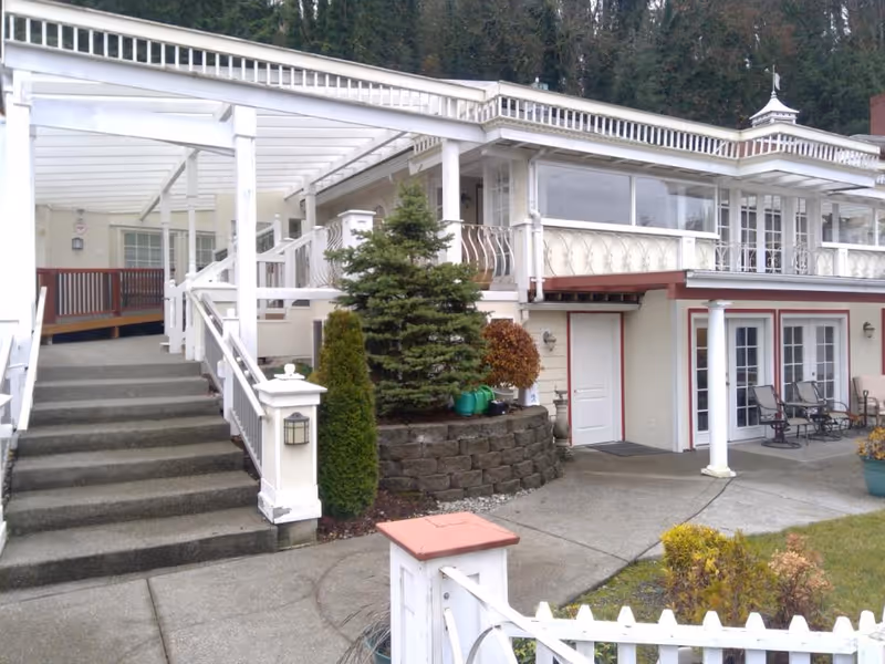 Exterior view of a two-story building with white railings and a covered porch area. There are stairs leading up to the porch, a small garden with shrubs and trees, and a concrete walkway. Patio chairs are visible near the lower level doors.