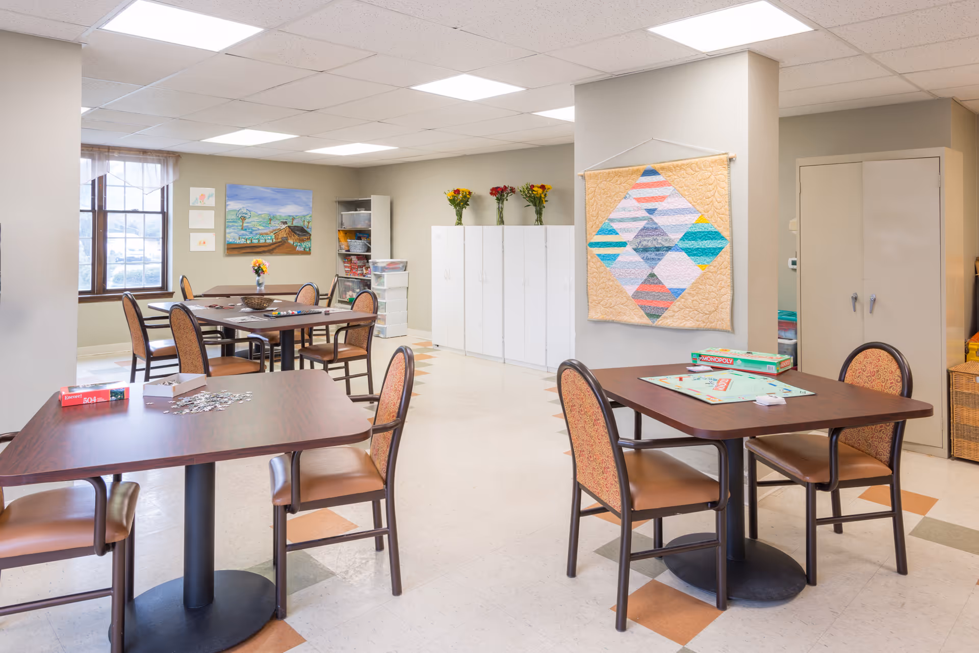 A bright, spacious activity room with several tables and chairs arranged for games and socializing. One table has a Monopoly board game set up, and another has a Scrabble game in progress. The room features a large window with sheer curtains, colorful artwork on the walls, a quilt hanging on a pillar, and white storage cabinets with vases of flowers on top.
