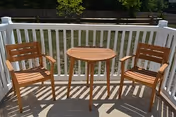 A small outdoor patio area with a round wooden table and two matching wooden chairs on a concrete floor, enclosed by a white railing with a grassy yard and trees visible in the background.