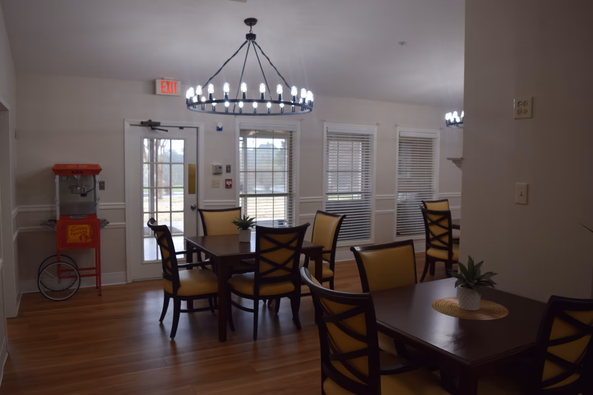 A senior living dining room with wooden tables, yellow-cushioned chairs, overhead chandeliers, windows, and a popcorn machine by the exit door.