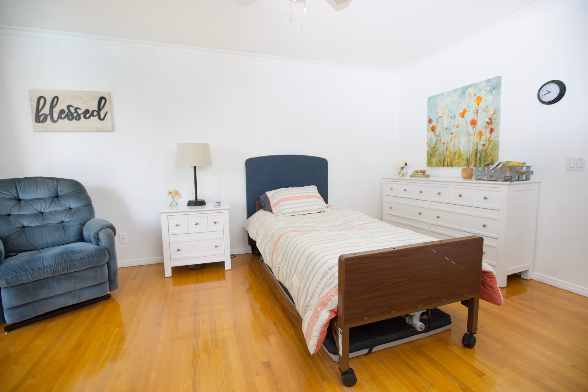 A bright and clean bedroom in a senior care facility featuring a single bed with a striped comforter, a blue upholstered armchair, a white nightstand with a lamp and small flower vase, a large white dresser with decorative items and a colorful floral painting above it, a wall clock, and a wooden floor. A wall sign reads 'blessed'.