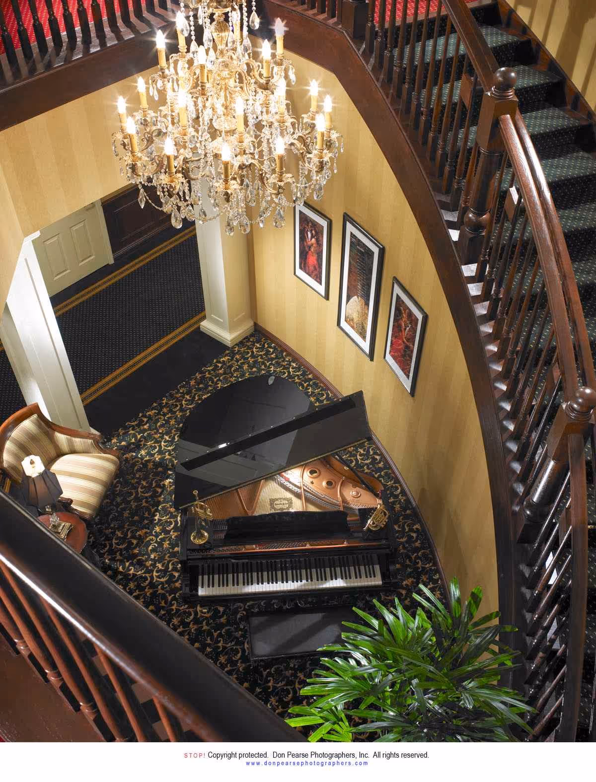 View from above of an elegant interior space featuring a grand piano on a patterned carpet, a striped armchair with a small side table and lamp, a large crystal chandelier hanging from the ceiling, three framed artworks on a yellow striped wall, and a wooden staircase curving around the area.
