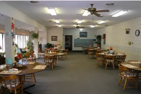 Communal dining room with multiple round wooden tables and chairs, plants, and ceiling fans.